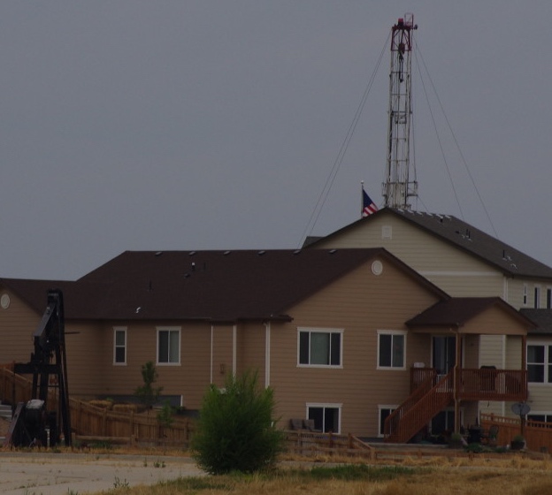 A pump jack sits in front of a house in northern Colorado. An drilling rig sits behind it. 