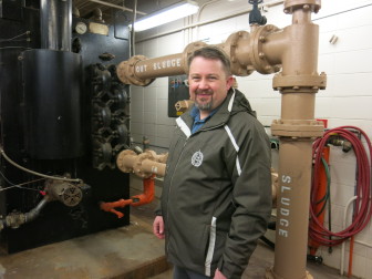 Clint Bassett stands in front of two thick pipes labeled "sludge in, sludge out."